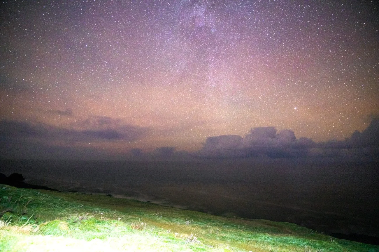 Ciel étoilé avec la Voie lactée visible au-dessus d'un champ verdoyant, avec des nuages sombres à l'horizon.