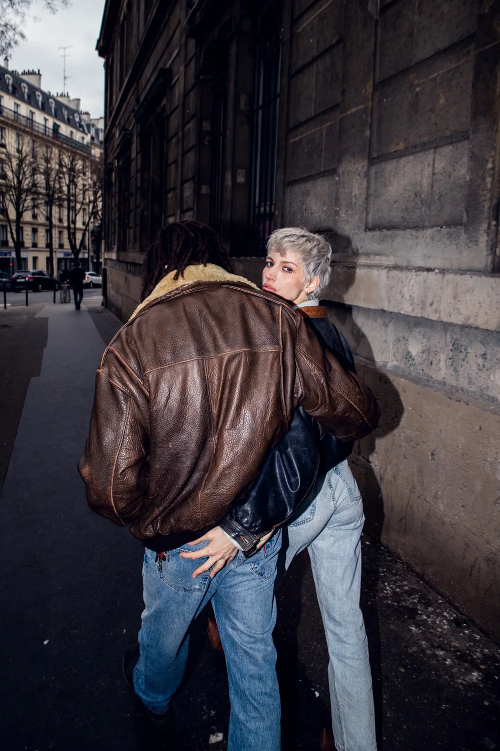 deux personnes debout contre un mur en pierre dans une rue urbaine, l'une regarde derrière elle avec les cheveux gris, l'autre avec des cheveux noirs porte un manteau en cuir brun.