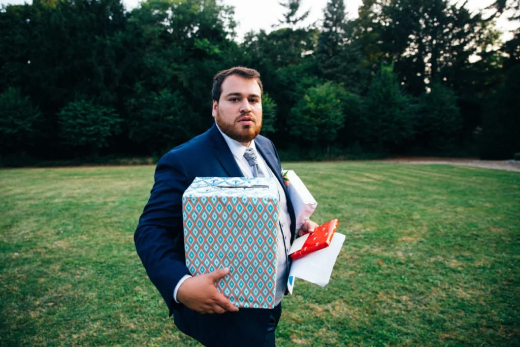 Un homme en costume tient un grand coffret cadeau et un bouquet de fleurs dans un espace extérieur vert.