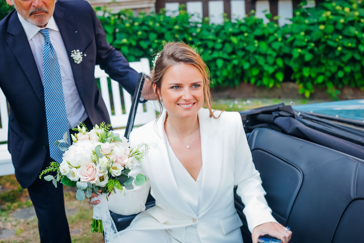 Une femme souriante en costume blanc tenant un bouquet de fleurs blanches, assise dans une voiture lors d'une occasion spéciale, probablement un mariage, avec un homme en costume derrière elle.