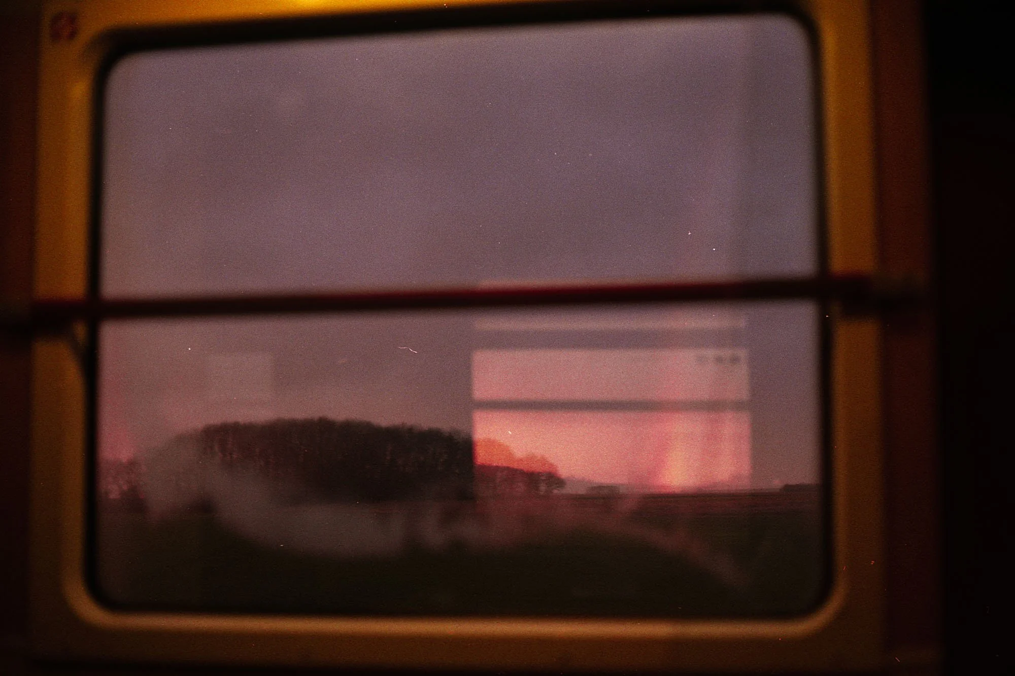 Coucher de soleil rougeâtre visible à travers une fenêtre de train ou de bus, avec silhouettes d'arbres et de collines à l'extérieur.