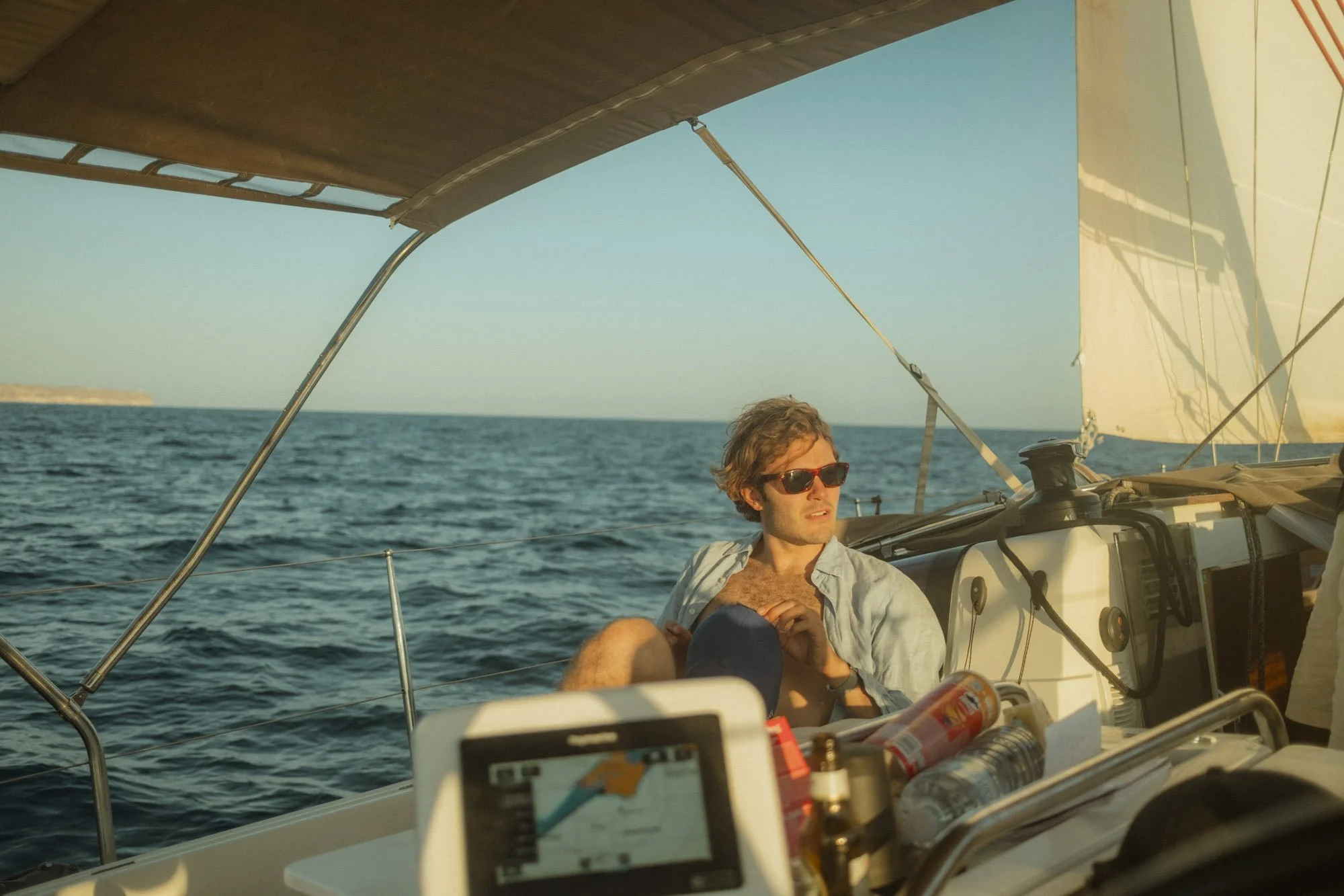 Un homme portant des lunettes de soleil assis sur un bateau à voile en pleine mer, avec des voiles blanches et un ciel clair à majorque