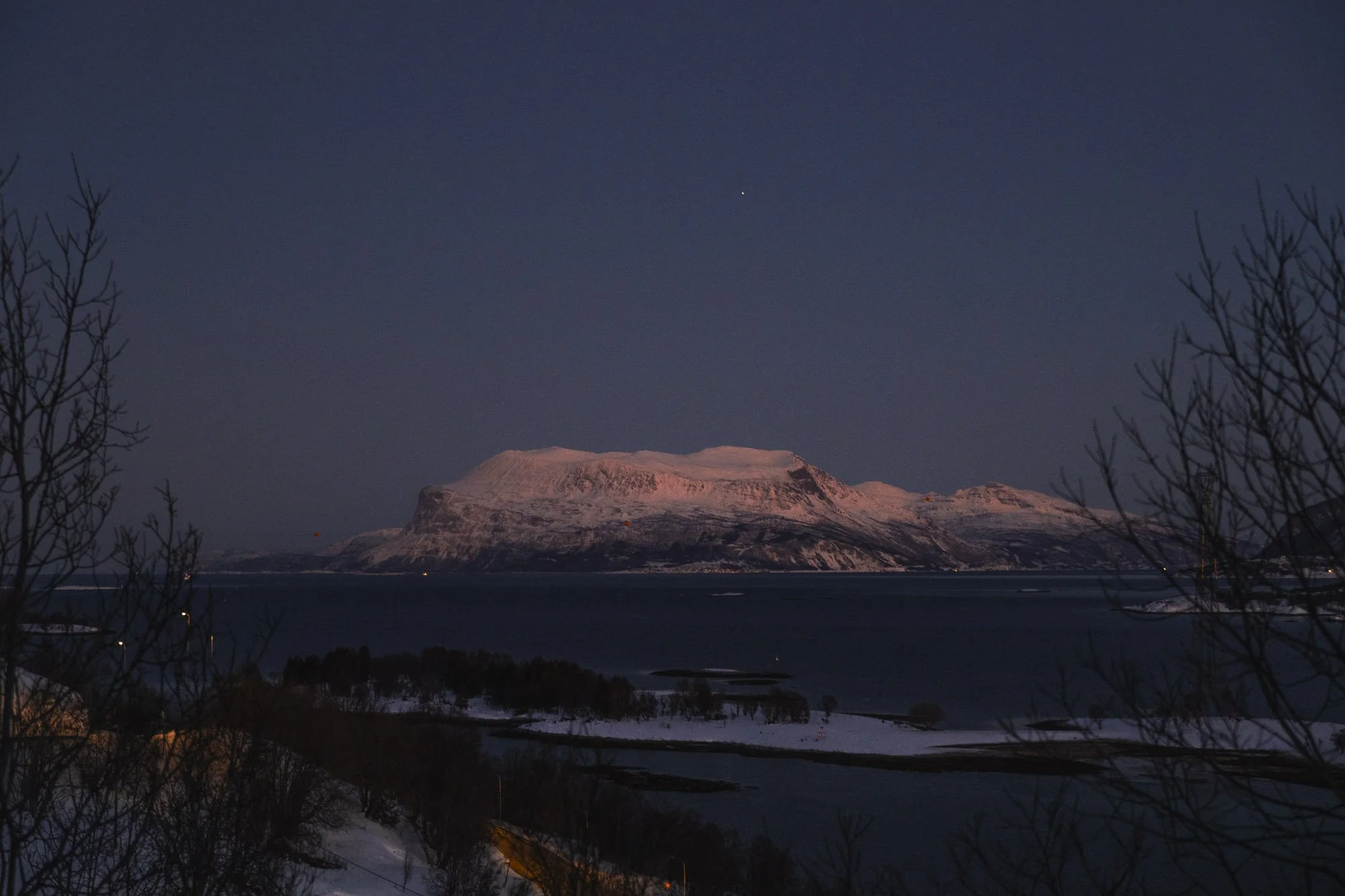 Une montagne enneigée au coucher du soleil, avec un ciel nocturne clair et une étoile visible, entourée de silhouettes d'arbres sans feuilles et d'une étendue d'eau froide à Tromsø.