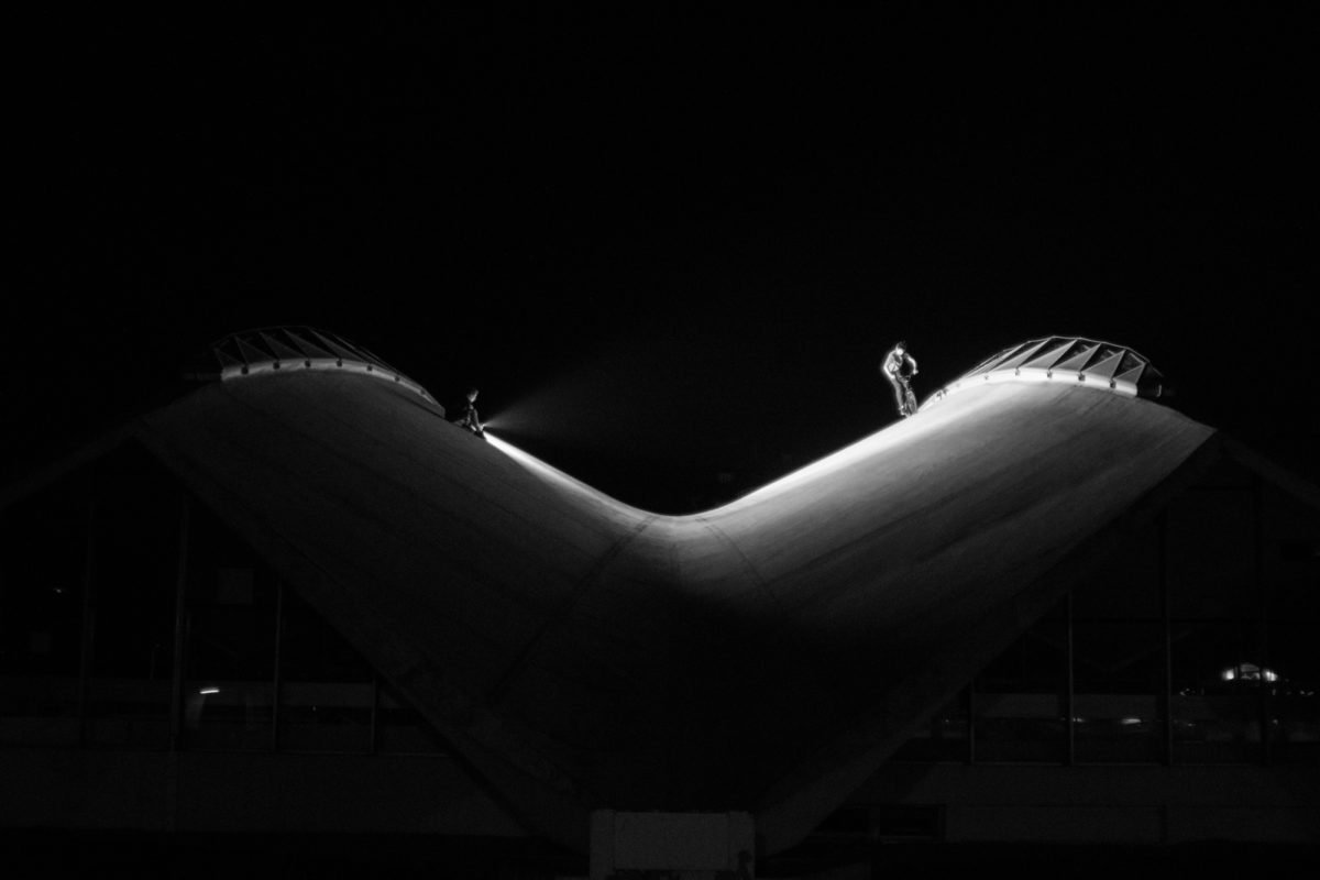 Deux personnes sur un toit en forme de vague, l'une avec une lampe de poche, en noir et blanc, la nuit.