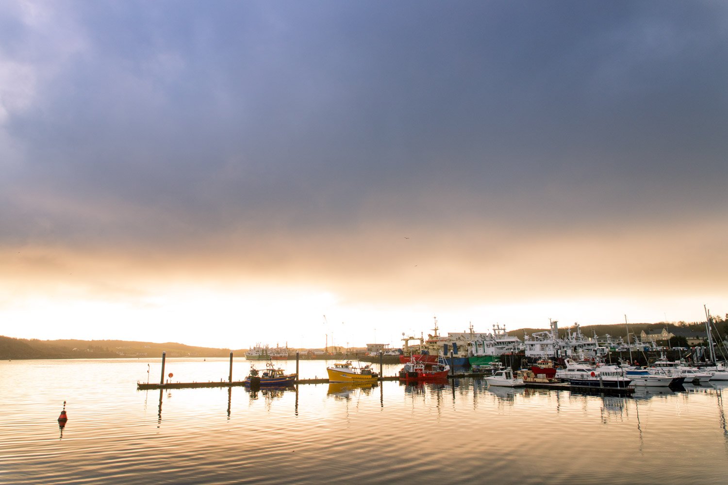 Port de pêche avec plusieurs bateaux amarrés, ciel sombre avec éclaircies au coucher du soleil