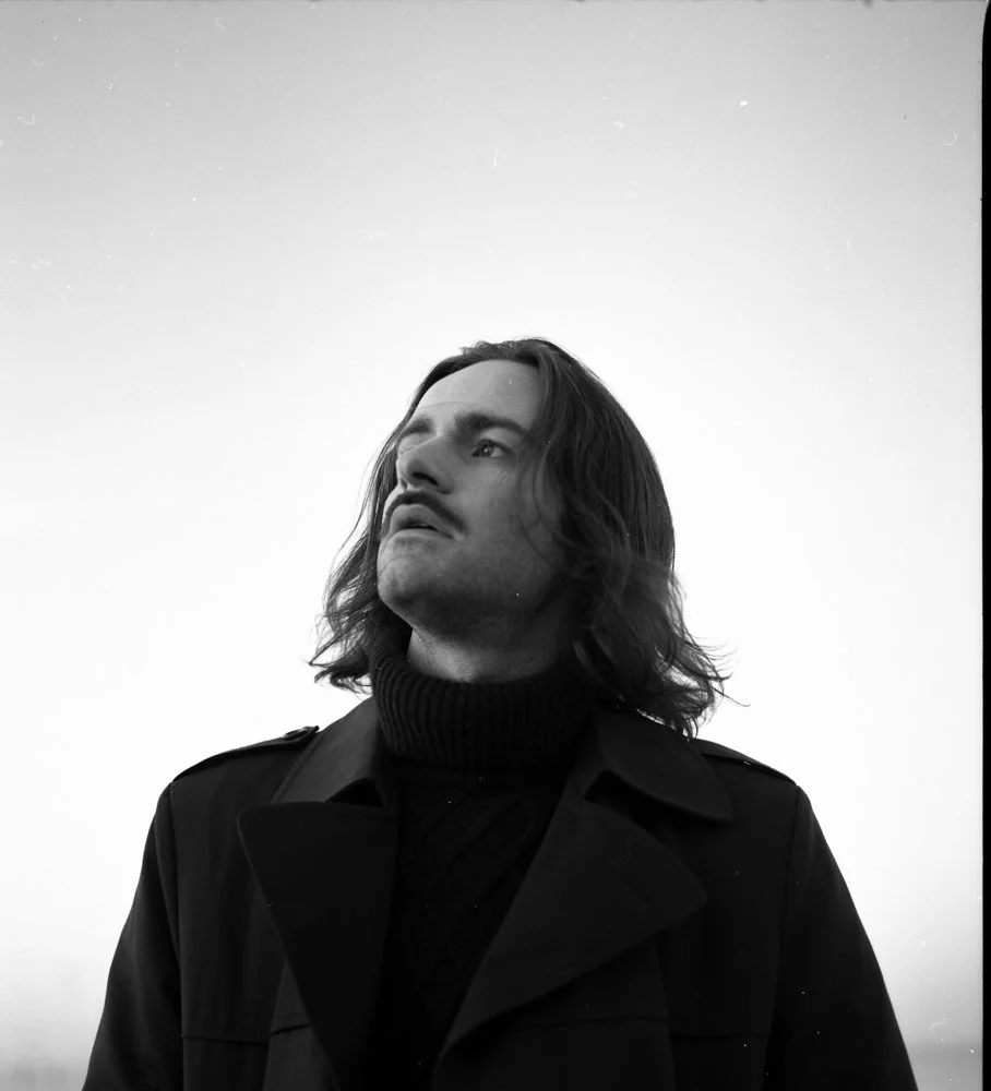 Un portrait d'un homme avec des cheveux longs portant un manteau noir, regardant vers la gauche, en noir et blanc.