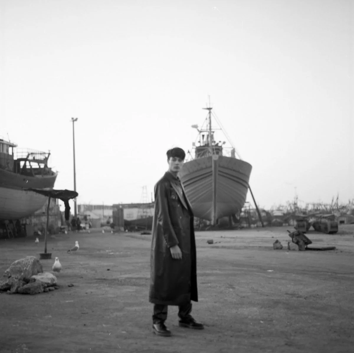 Un jeune homme en manteau long se tient dans un port avec des bateaux déserts en arrière-plan, en noir et blanc.