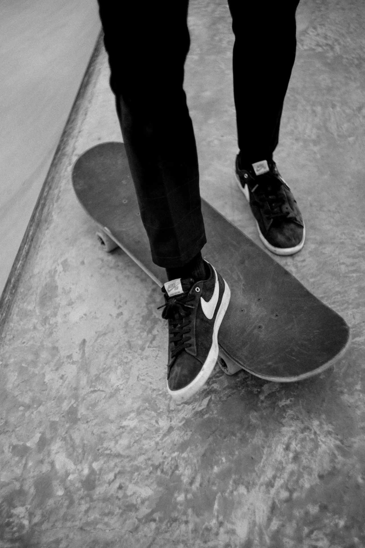 Personne debout sur un skateboard dans une pièce avec sol en béton, portant des chaussures de sport Nike et un pantalon noir.