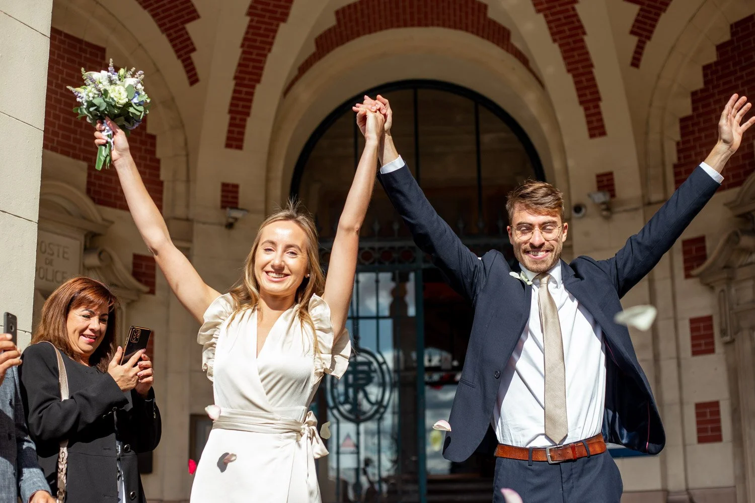 Un couple heureux, jeune femme en robe claire et jeune homme en costume, célébrant leur mariage devant un bâtiment en pierre avec une arche en arrière-plan, de la fête et des invités qui prennent des photos autour d'eux.