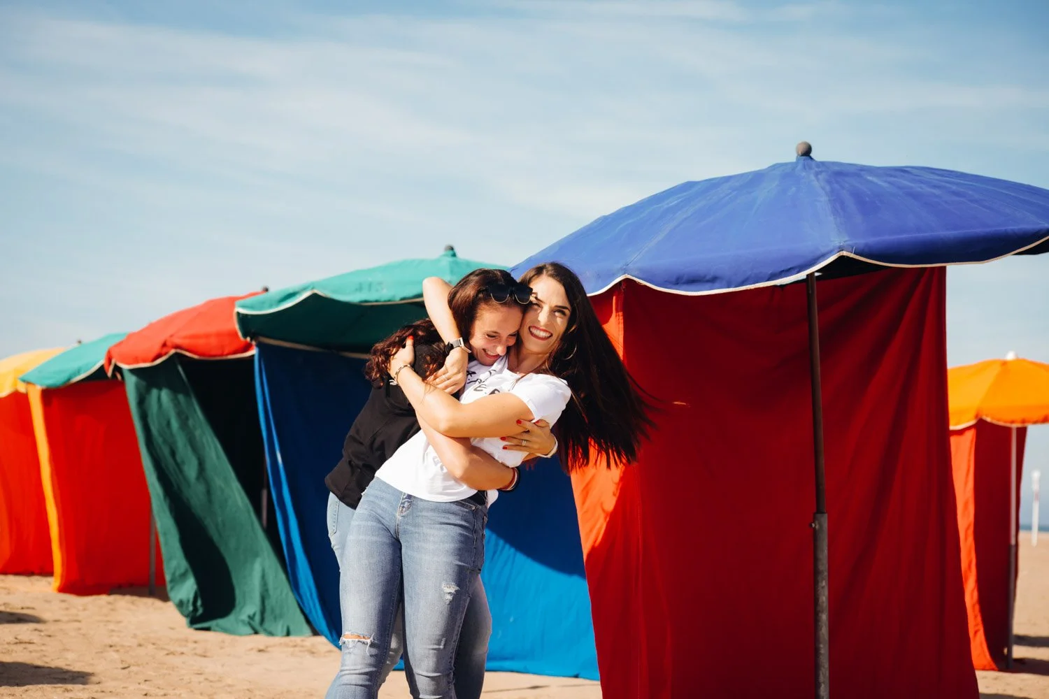Deux femmes souriantes s'embrassent sur une plage avec des cabines de plage colorées en arrière-plan.