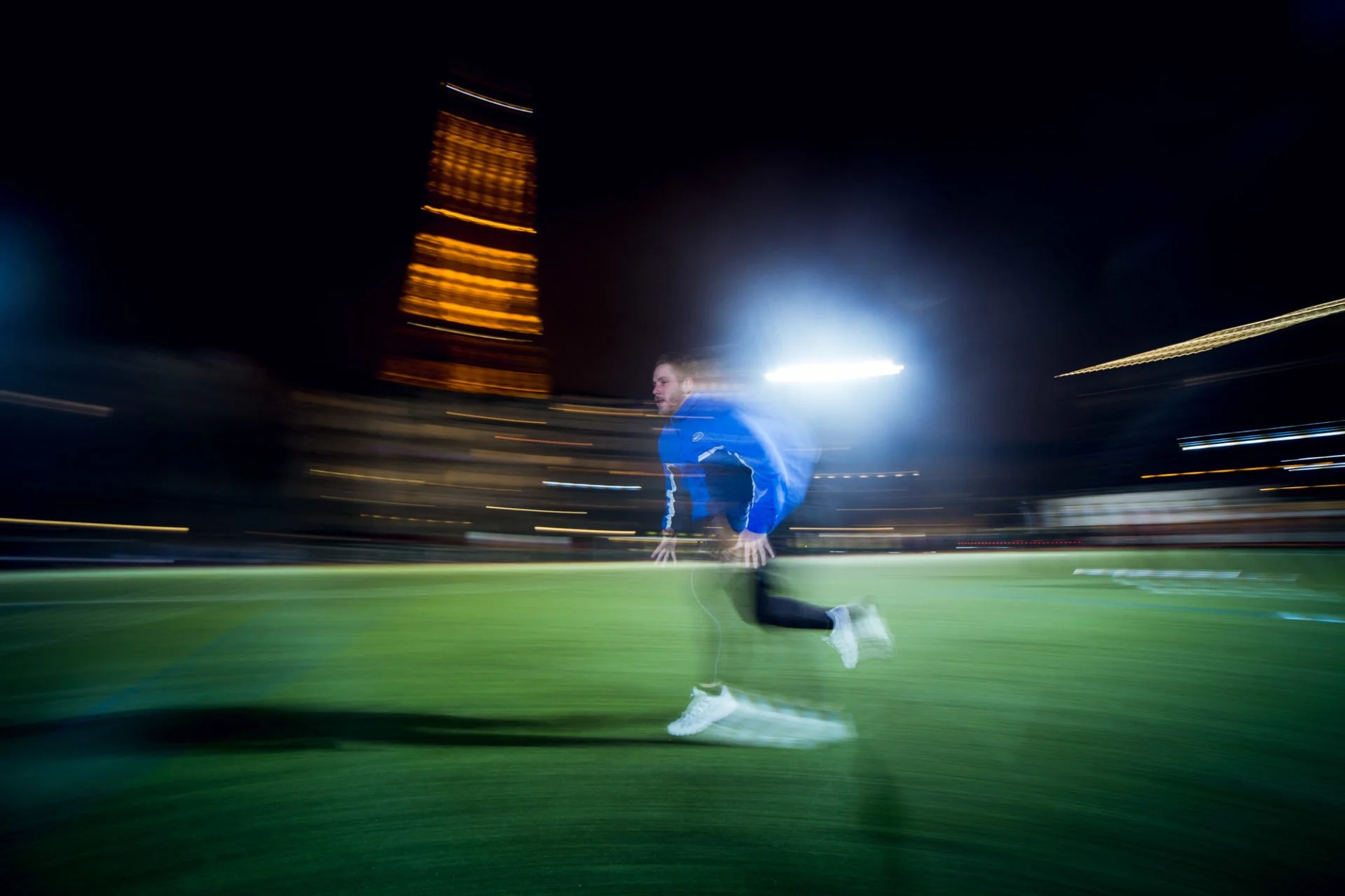 Paul Deby en mouvement rapide s'entraîne ou court sur un terrain de sport la nuit, avec un immeuble illuminé en arrière-plan et des effets de lumière laissant des traces de vitesse.