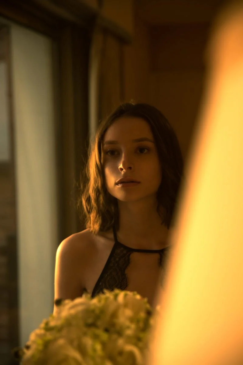Une jeune femme regarde son reflet dans un miroir dans une pièce chaleureuse, éclairée par une lumière douce.