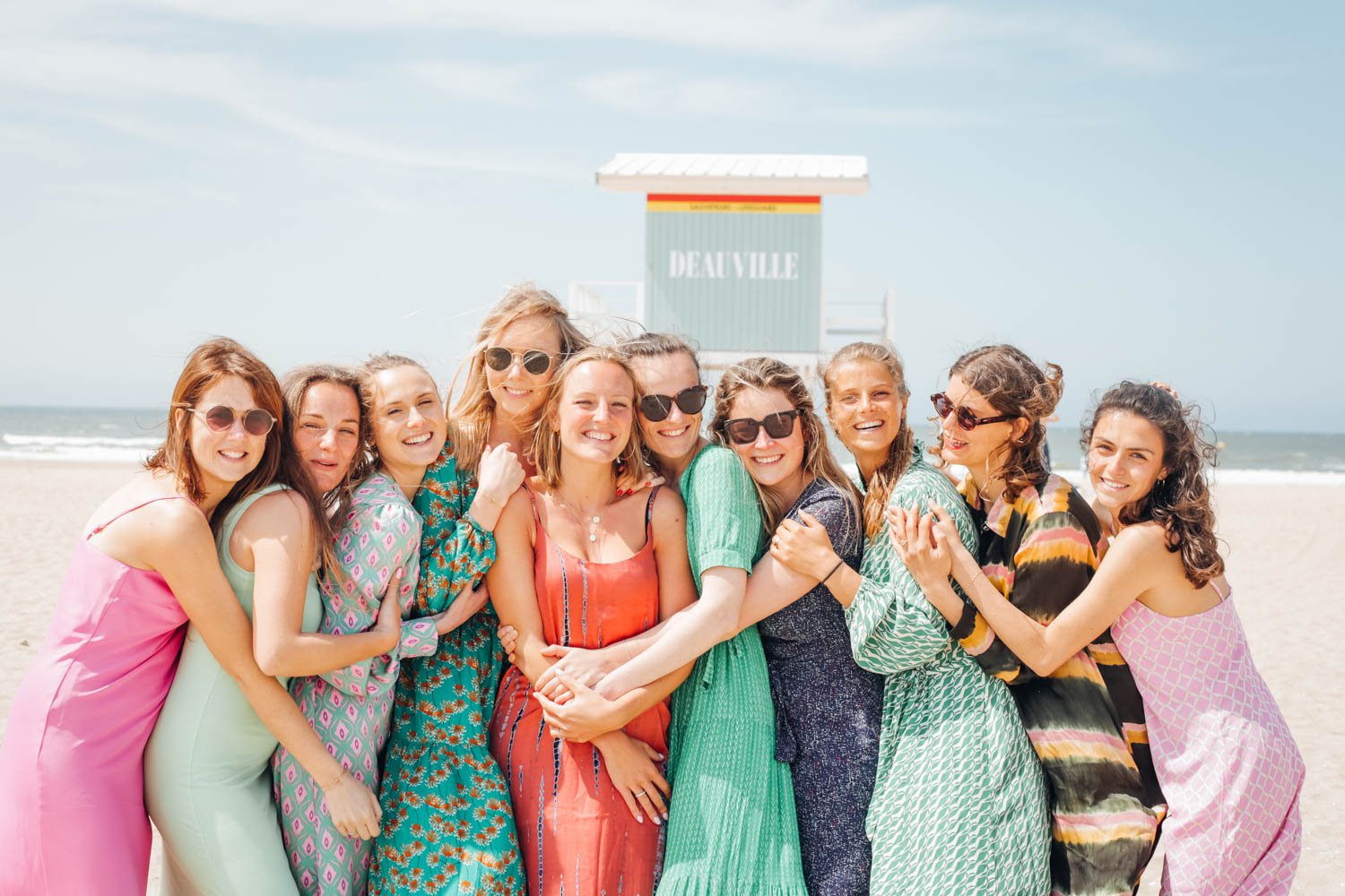 Groupe de femmes souriantes portant des robes colorées sur une plage, avec le ciel bleu et la mer en arrière-plan.