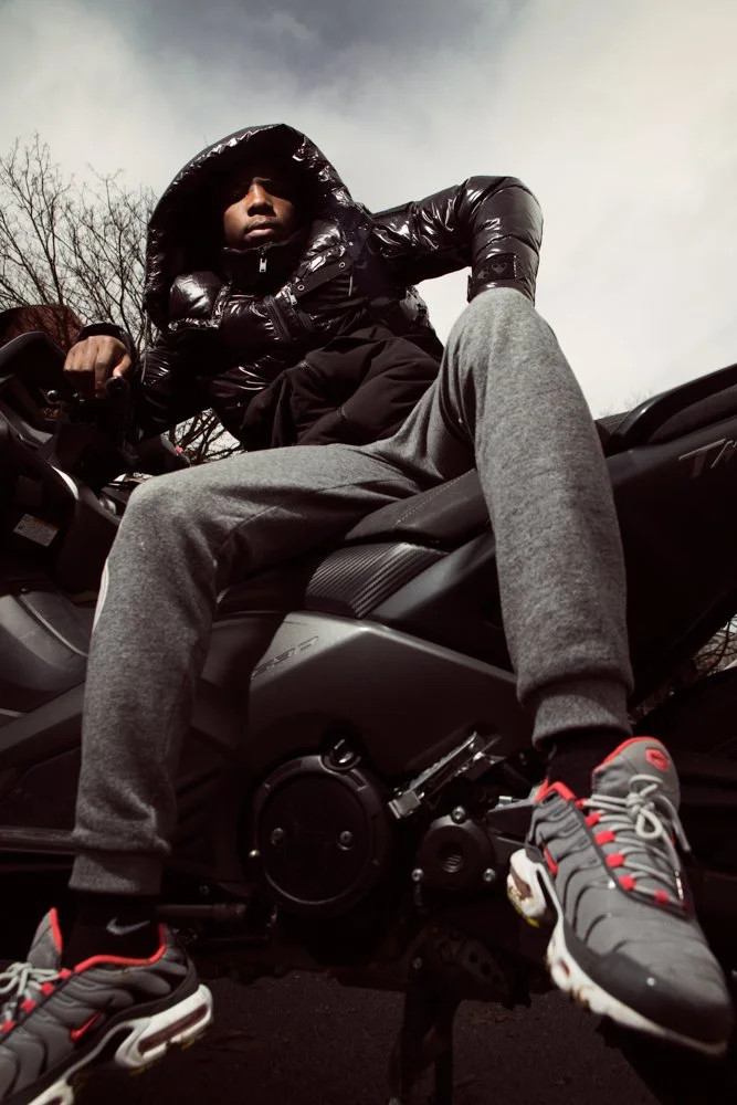 Une personne portant une veste en cuir noire, un capuchon, des pantalons gris et des chaussures de sport grise, rouge, noire, s'assoit sur une moto, vue de dessous, arrière-plan avec ciel gris et arbres dénudés.