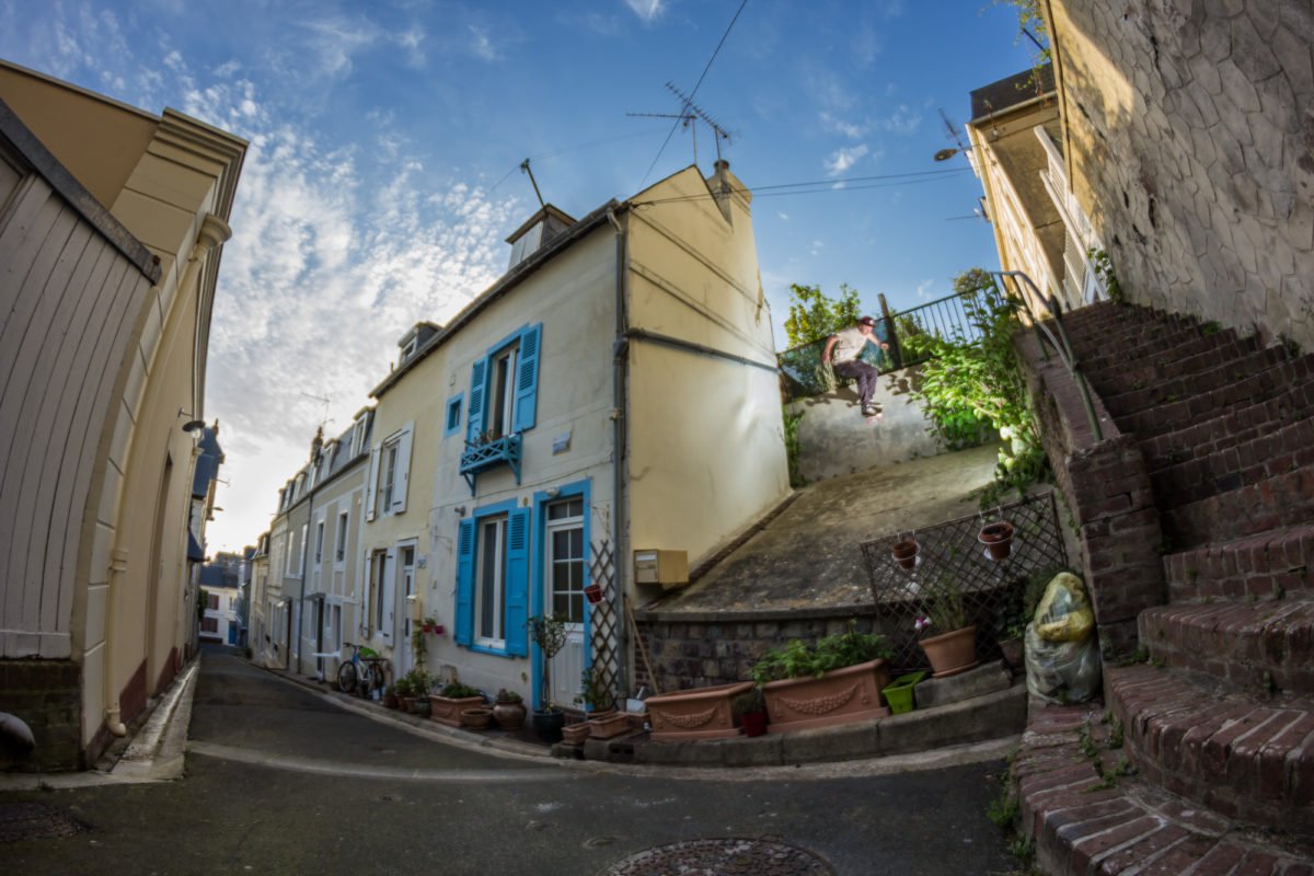 Rue étroite avec des maisons peintes en blanc et des volets bleus. Un homme s'assoit sur une clôture en haut d'une pente escarpée. Il y a des plantes en pot devant les maisons et des escaliers en briques à droite.