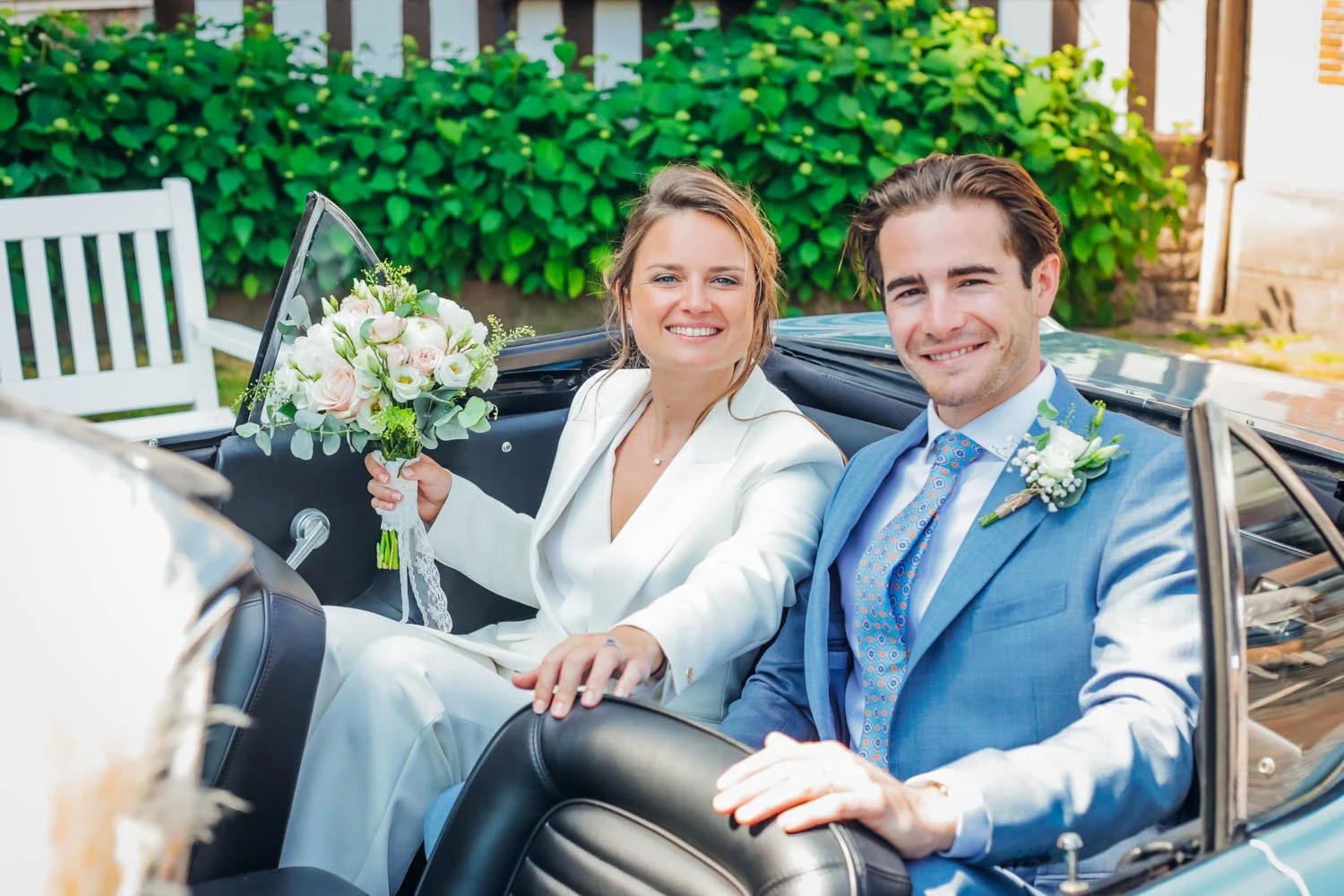 Un couple de mariés souriants en voiture décapotable : la femme en costume blanc et le homme en costume bleu avec cravate, portant des fleurs de mariage.