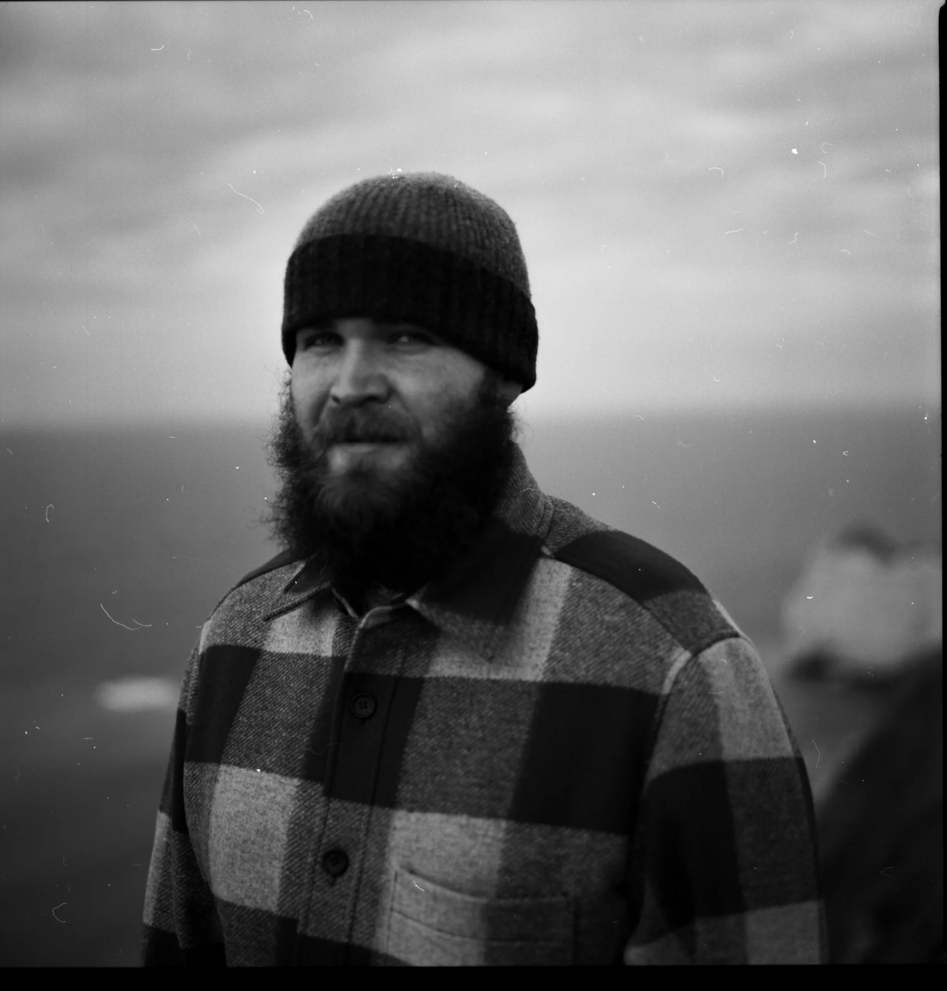 Un homme portant un bonnet en laine et un pull à carreaux, avec une barbe, regardant vers la caméra, avec un ciel nuageux en arrière-plan à étretar