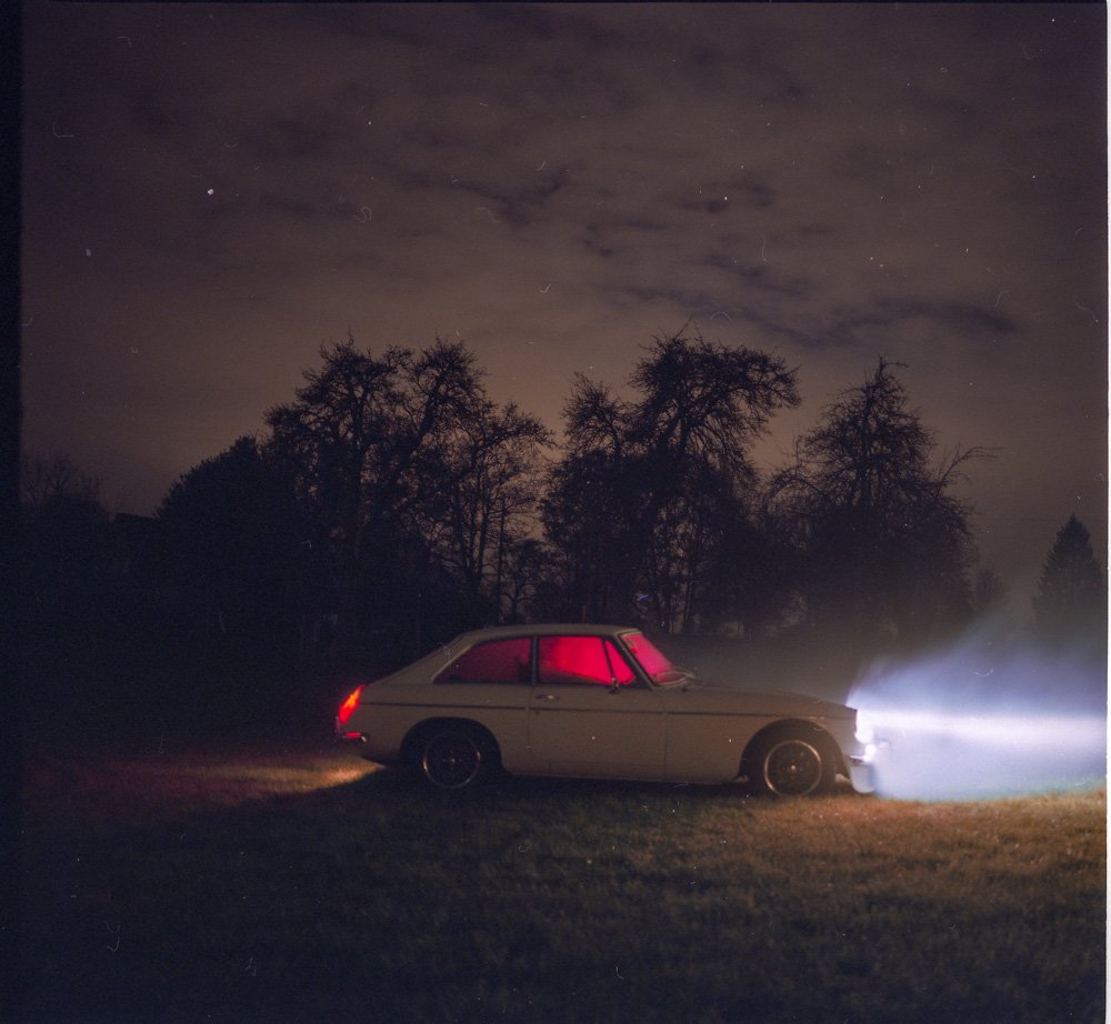 Une voiture ancienne blanche avec un intérieur rouge, garée sur une pelouse sombre, avec des phares allumés projetant de la lumière dans l'obscurité, sous un ciel nuageux et des arbres dénudés.