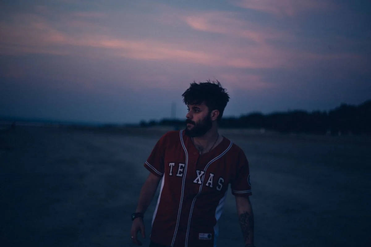 Jeune homme avec barbe et tatouages, portant un maillot de baseball "TEXAS", se tenant à l'extérieur au coucher du soleil, avec un ciel coloré en arrière-plan.
