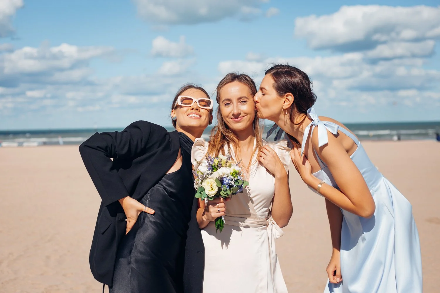 Trois femmes sur la plage, l'une en robe blanche tenant un bouquet de fleurs, une autre en costume noir avec des lunettes blanches, et la troisième en robe bleue donnant un baiser sur la joue à la femme en blanc.