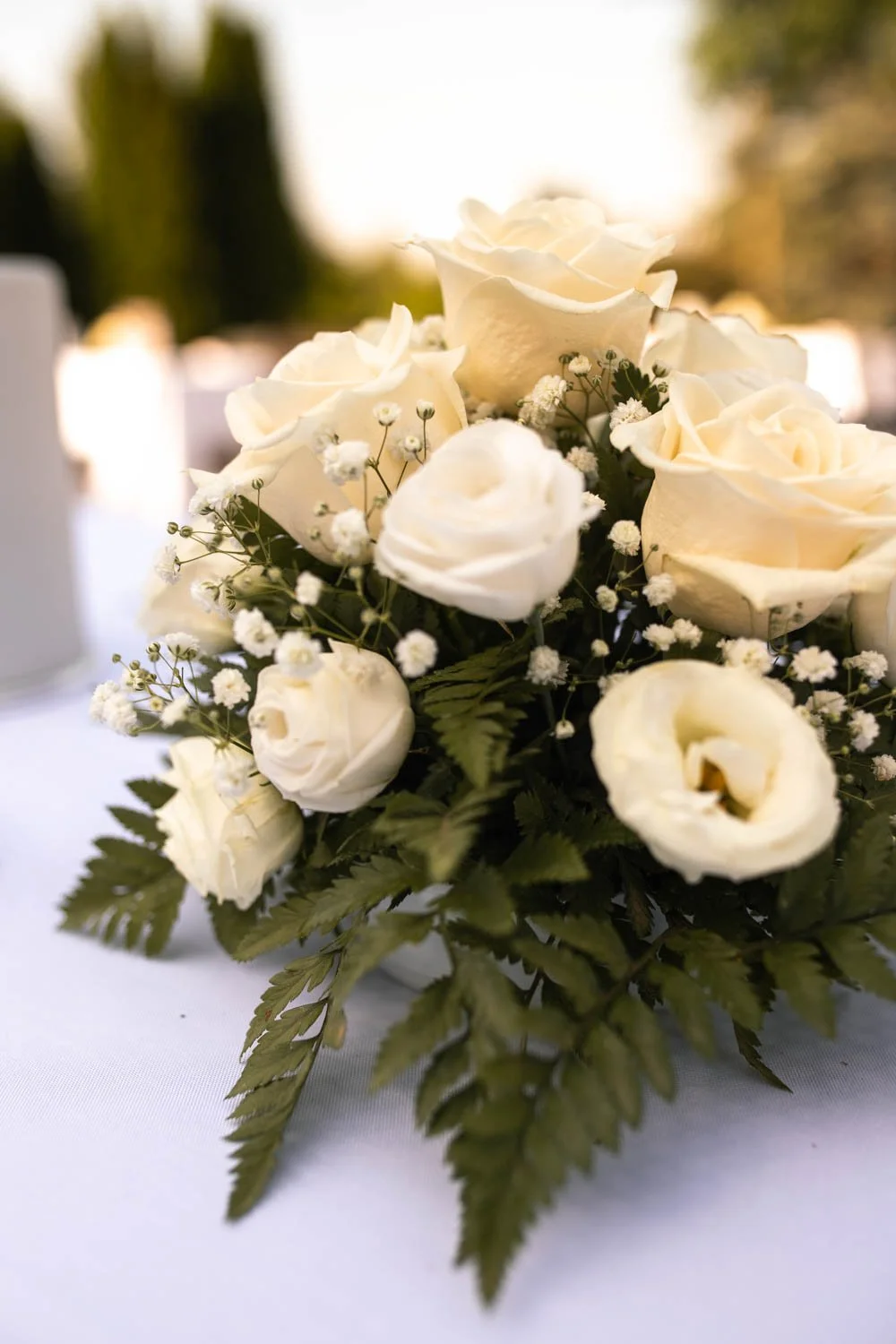 Un bouquet de roses blanches et petites fleurs blanches, entouré de feuillage vert, posé sur une table lors d'un événement en plein air.