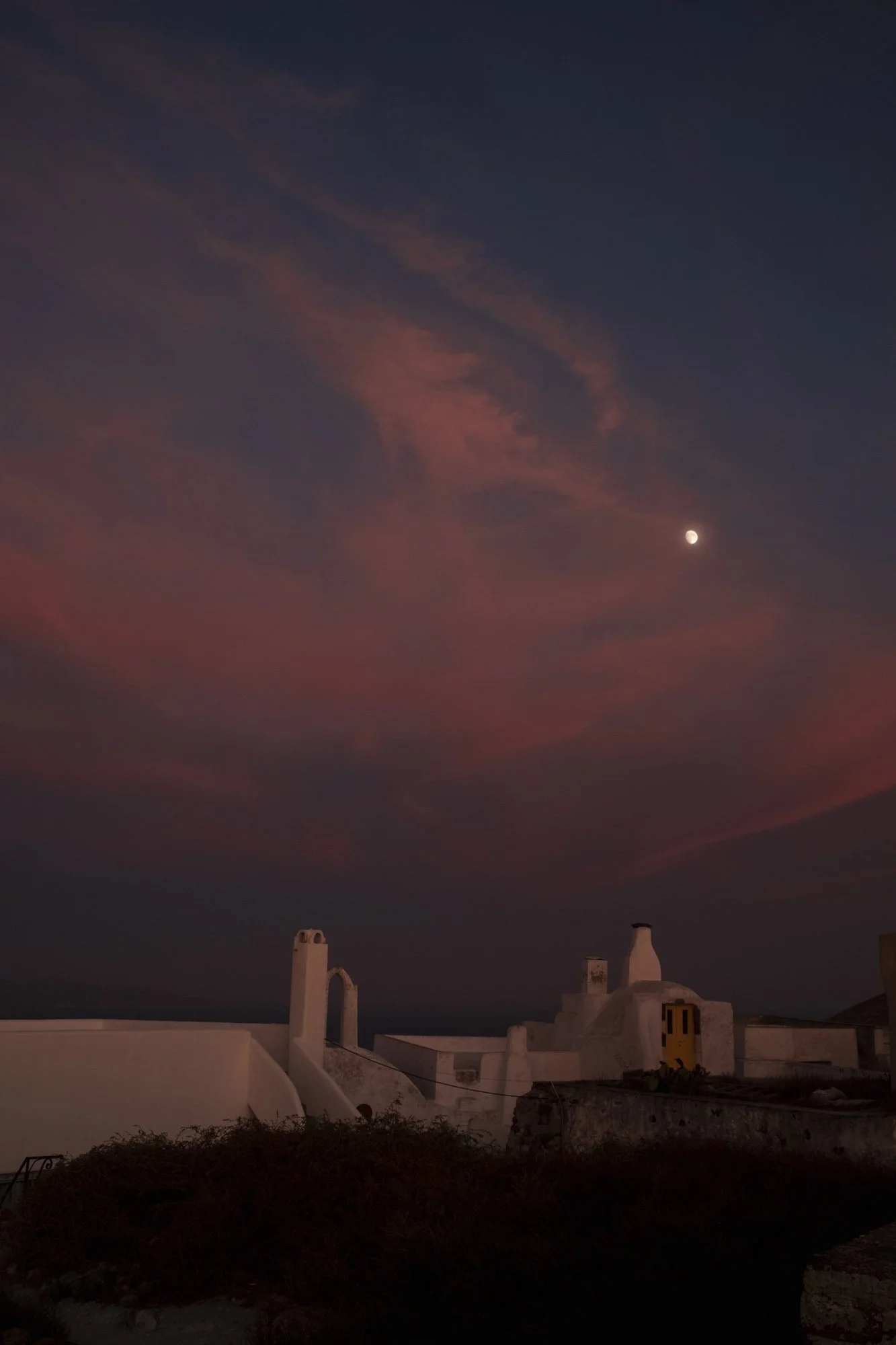 Ciel nocturne avec la lune visible et quelques nuages roses, bâtiments blancs en façade au premier plan, ambiance calme et pittoresque à santorin