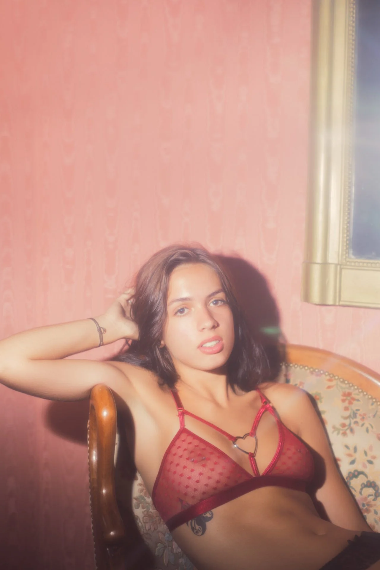 Une jeune femme aux cheveux bruns, portant un lingerie rouge en dentelle transparente, est assise sur une chaise avec un fond de mur rose et un cadre doré. Elle pose avec un bras derrière la tête, regardant l'objectif.