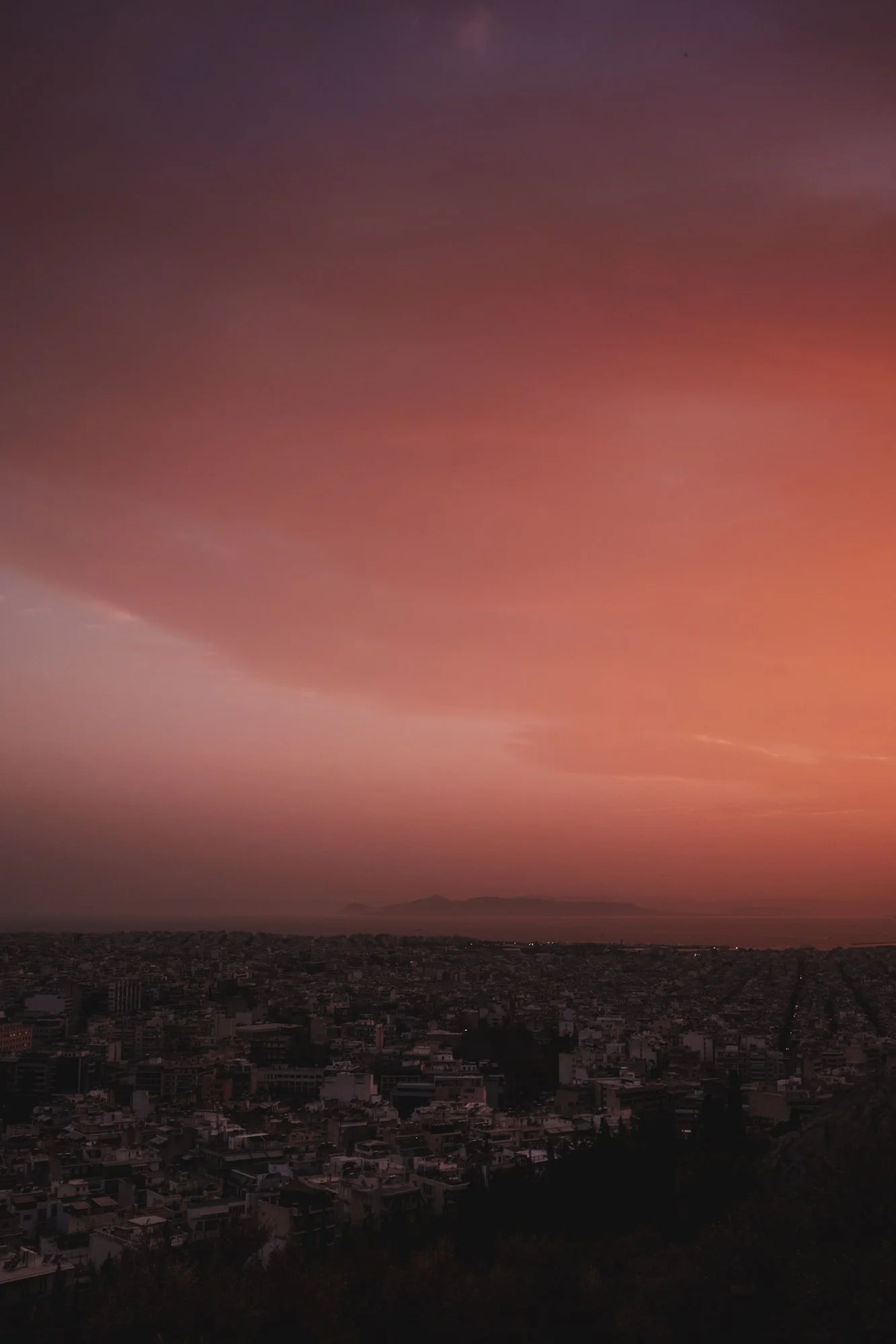 Vue urbaine au coucher du soleil avec un ciel rose-orange, une ville dense avec des bâtiments et des toits, et une montagne ou un volcan au loin à athenes