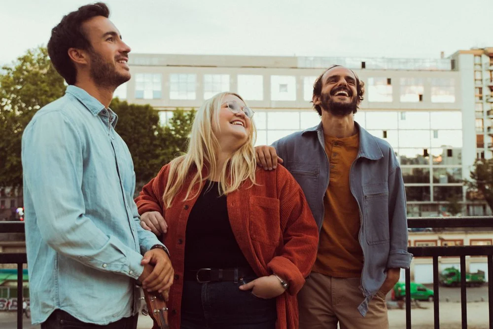 Trois jeunes adultes souriants et rigolant ensemble dans un espace extérieur urbain, l'un d'eux porte une veste en jean, une femme porte un manteau orange et un homme porte un blouson en jean.
