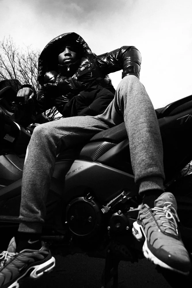 Un jeune homme en veste en plastique noir, capuche sur la tête, pose sur une moto en noir et blanc, vu de dessous, avec un ciel nuageux en arrière-plan.