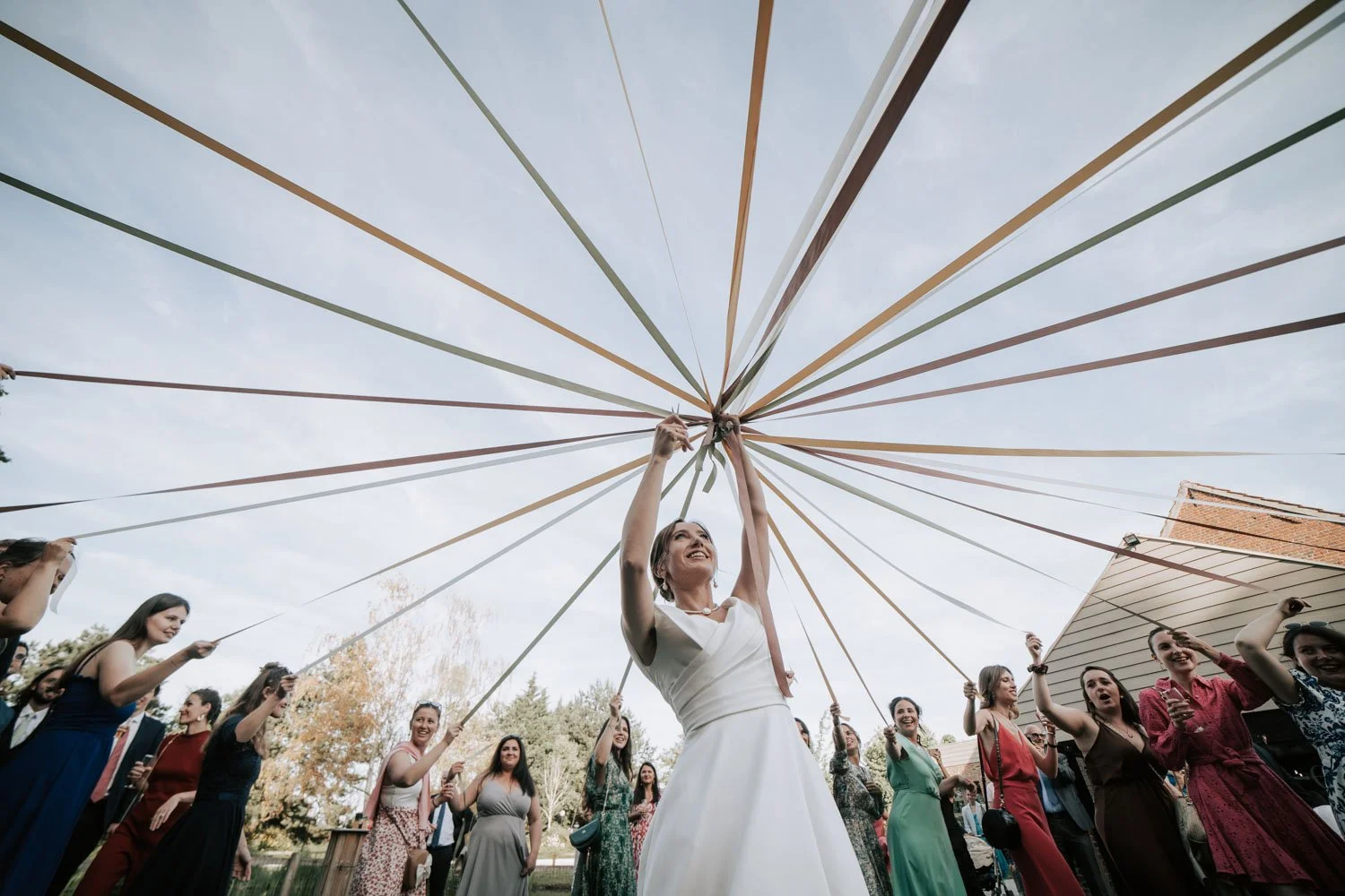 Groupe de personnes lors d'une fête d'extérieur, la mariée en robe blanche au centre, tenant une corde à l'envers avec un sourire, entourée d'invités en vêtements colorés, sous un ciel clair avec un parasol