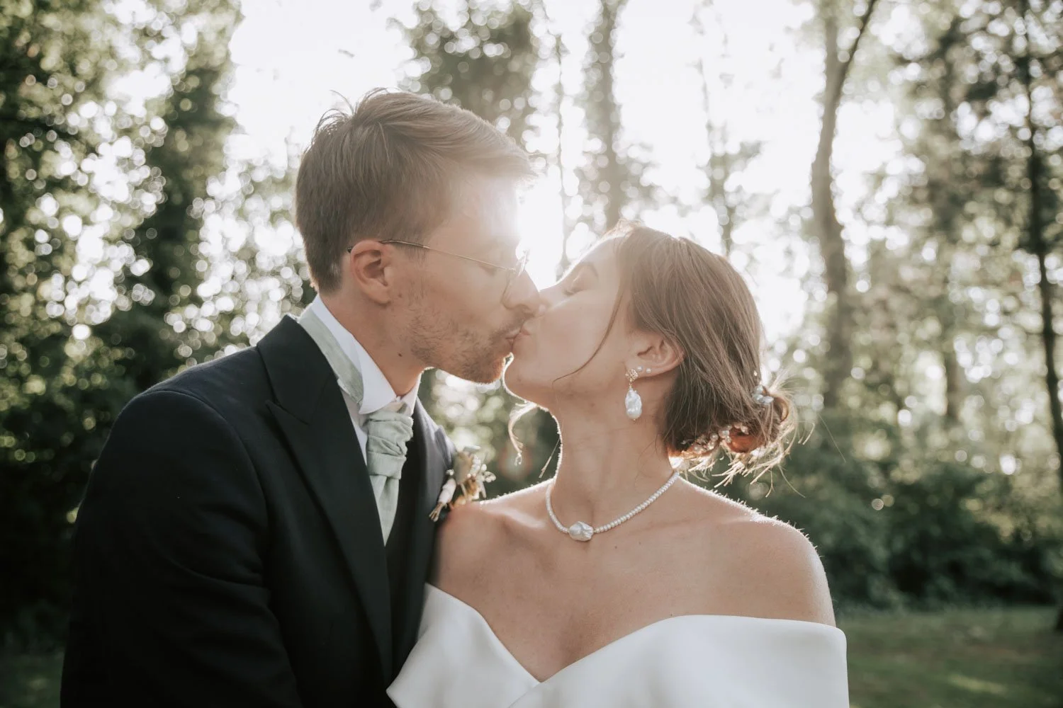 Un couple de mariés s'embrassant dans une forêt, avec la lumière du soleil derrière eux, lors d'un mariage en plein air.