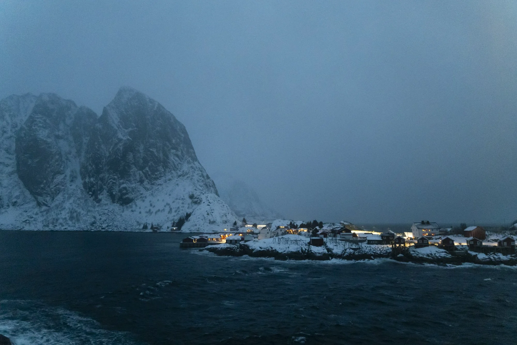 Village de pêcheurs au bord de la mer à tromso, avec montagnes enneigées en arrière-plan, lors d'une soirée ou matinée sombre.