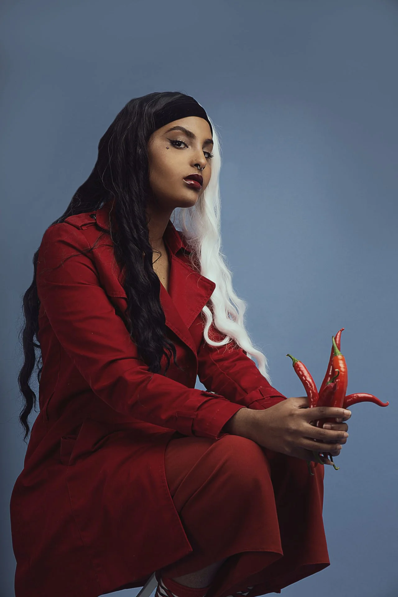 Une femme avec des cheveux mi-noirs, mi-blancs, portant une veste rouge, assise contre un fond bleu, tenant un groupe de piments rouges dans ses mains.