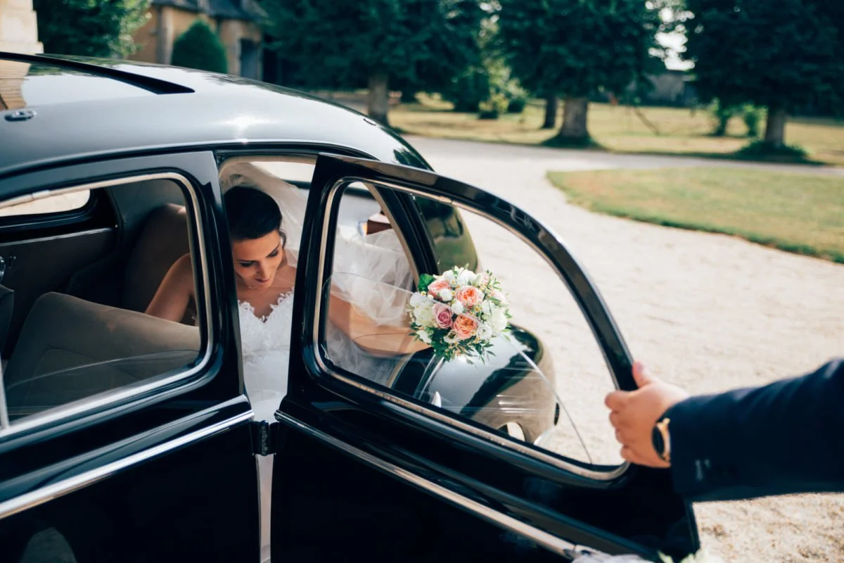 Une mariée sort d'une voiture noire de charme, tenant un bouquet de fleurs, pendant qu'une personne ouvre la porte. Le décor extérieur est composé d'arbres et d'une allée.