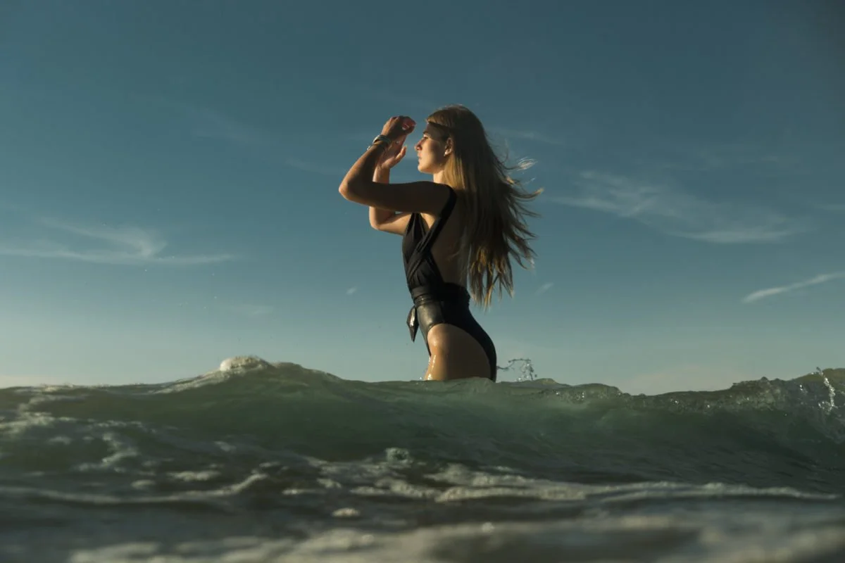Une femme en maillot de bain regarde au loin en tenant ses mains en visière dans la mer, sous un ciel clair.