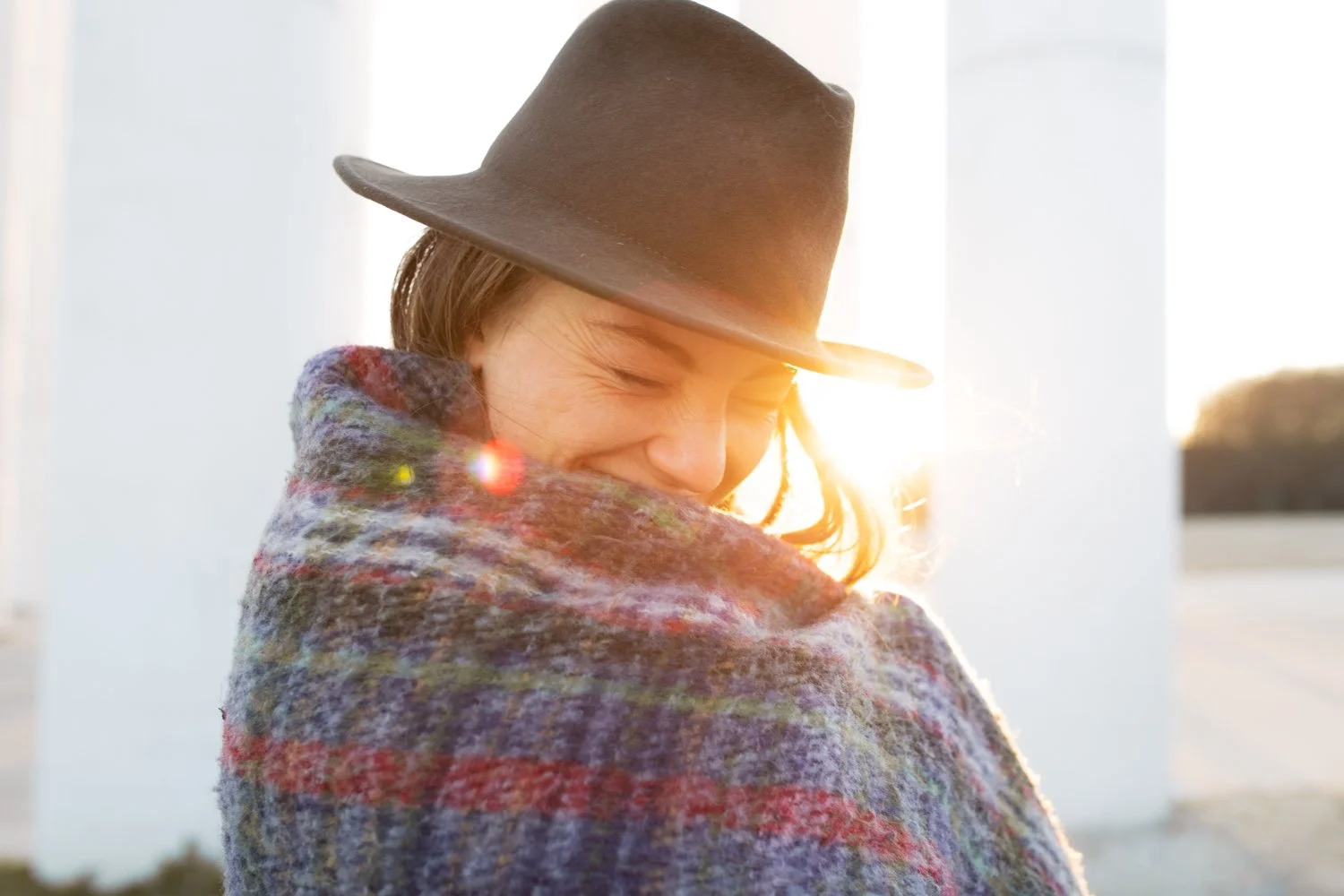 Une femme souriante, portant un chapeau en terre cuite, enroulée dans une écharpe colorée, avec le soleil couchant en arrière-plan.