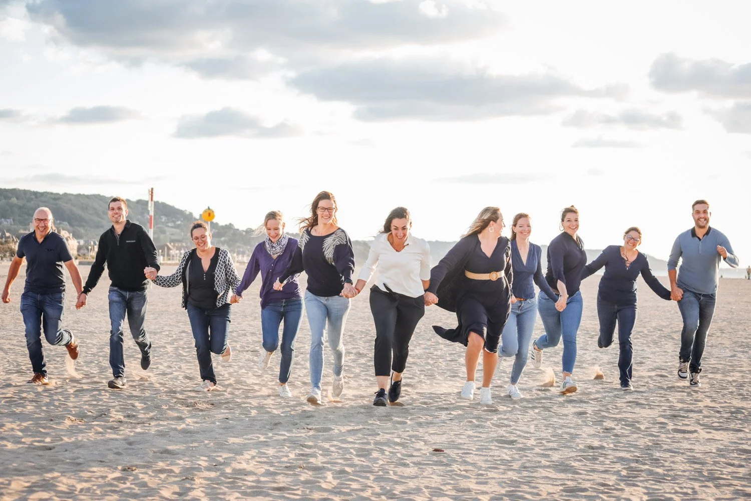 Groupe de dix personnes souriantes qui marche ensemble sur la plage par une journée ensoleillée, tenant la main.
