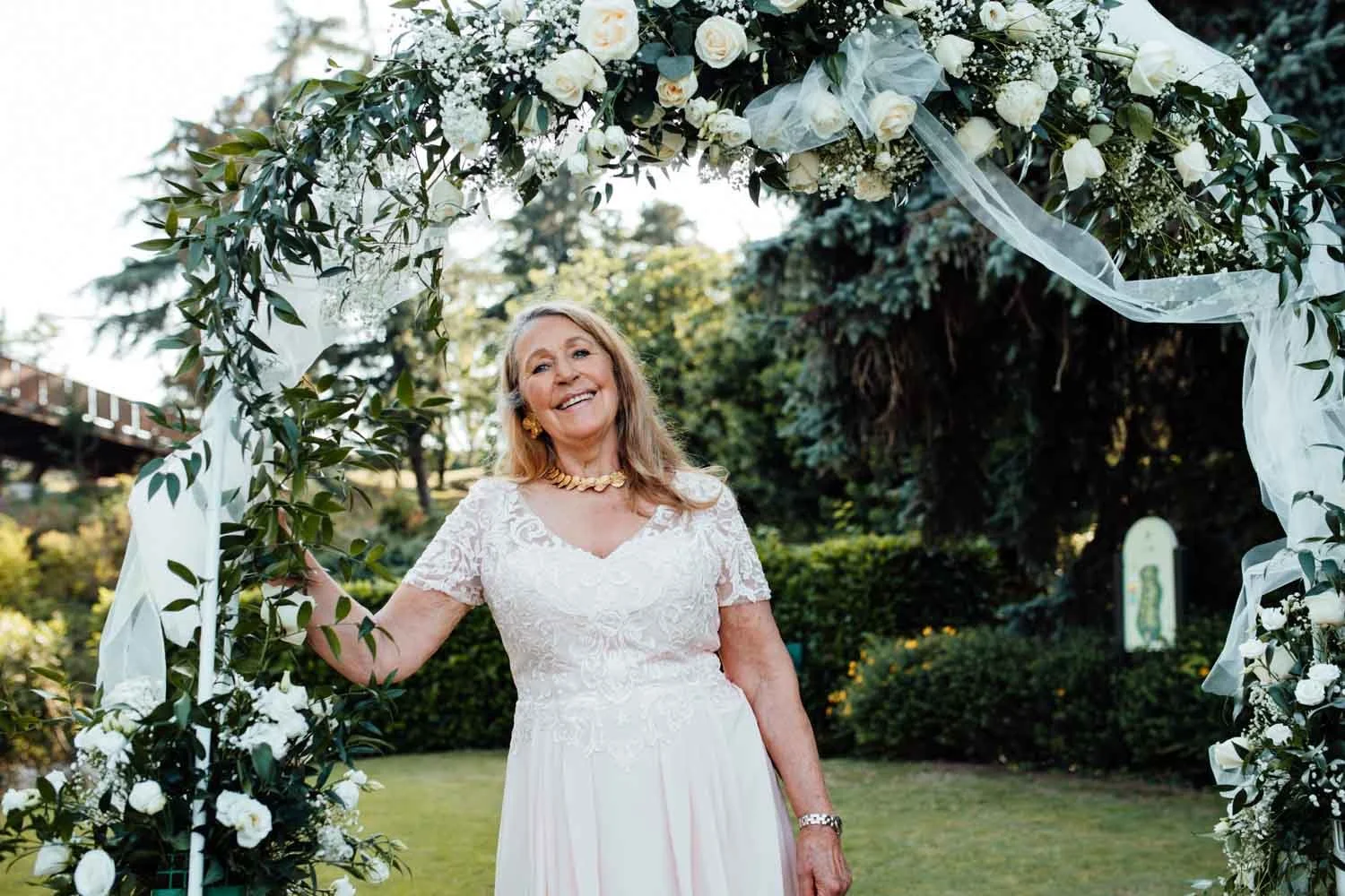 Une femme âgée en robe blanche élégante, portant un collier et des boucles d'oreilles dorés, souriante, debout sous une arche décorative faite de fleurs blanches et de verdure lors d'une cérémonie en extérieur.