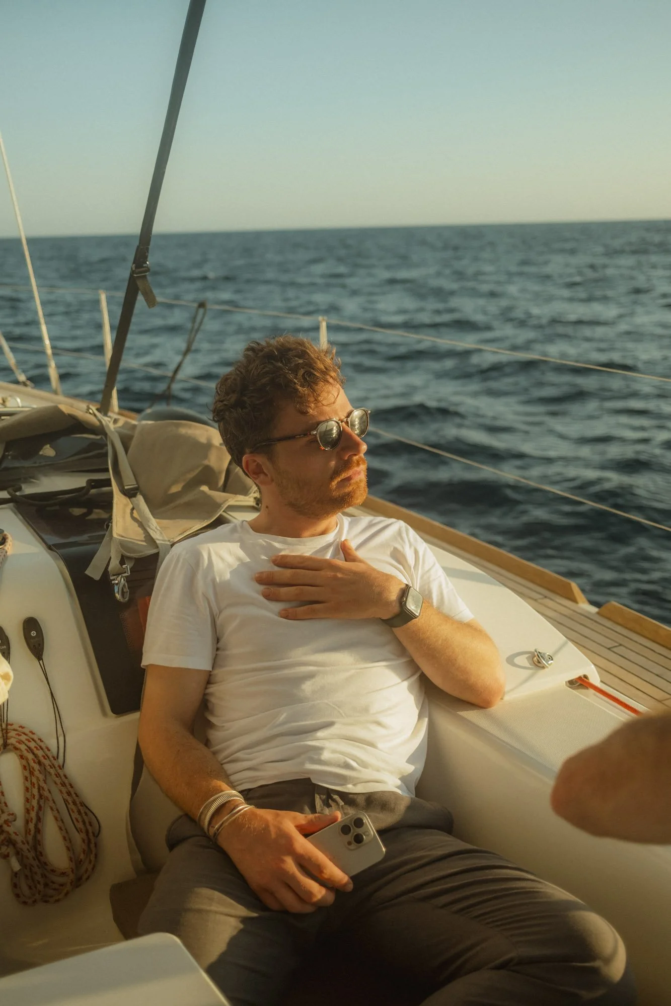 Un homme avec des lunettes de soleil en blanc t-shirt, détendu sur un bateau en mer, tenant un téléphone portable. Le ciel est clair et la mer est calme à majorque