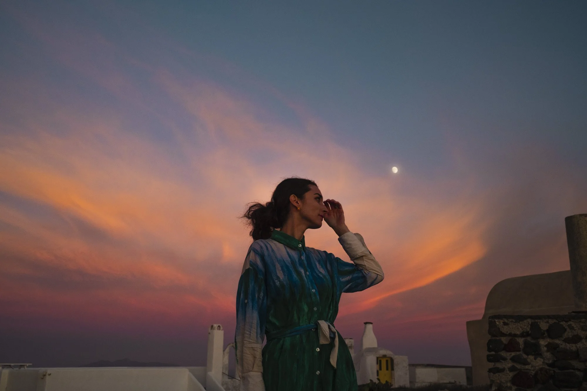 Une femme regarde le ciel au coucher du soleil, avec une couleur rose et orange, en portant un manteau à rayures vertes et bleues, face à la mer ou à l'océan et un ciel étoilé avec la lune visible à santorin