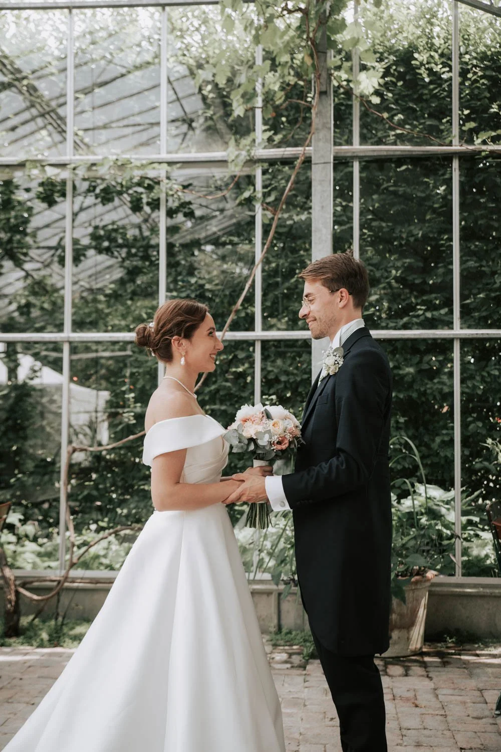 Un couple de mariés échange des vœux dans une serre, la femme en robe blanche avec une coiffure en chignon, l'homme en costume noir, tenant un bouquet de fleurs.