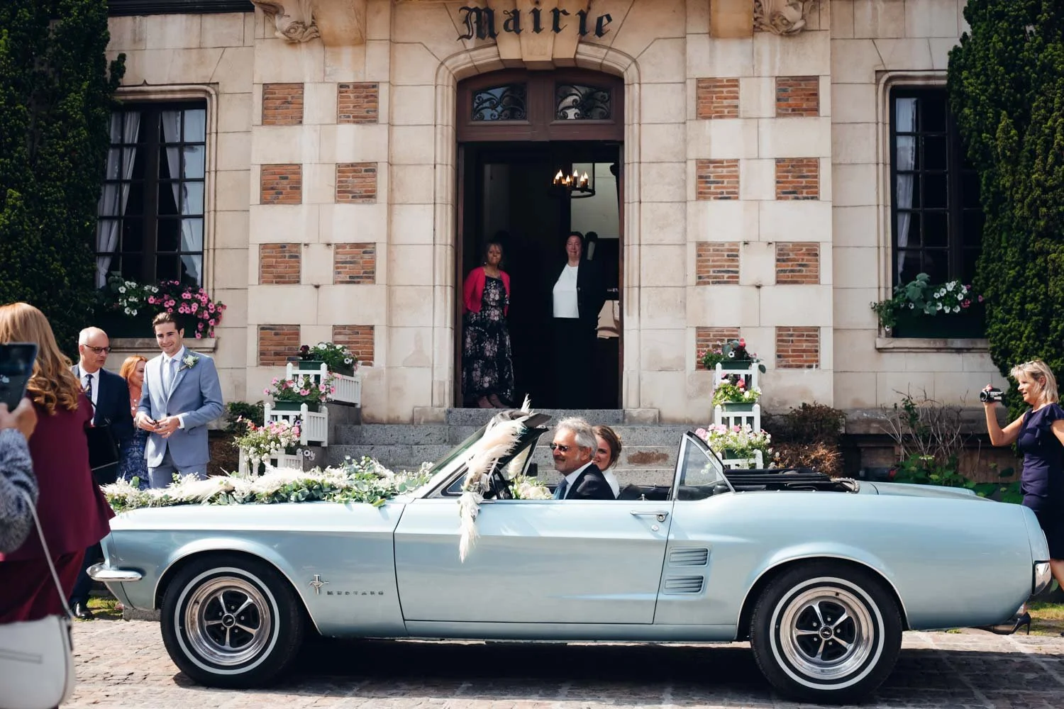 Voiture de mariage vintage bleue garée devant une église, avec deux personnes à l'intérieur et des invités en arrière-plan. L'église porte une enseigne 'Mairie' au-dessus de l'entrée.