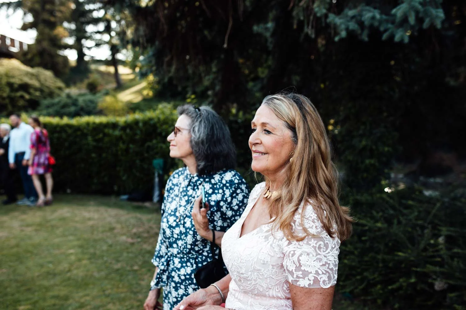 Deux femmes debout dans un jardin, souriantes, lors d'un événement en plein air, avec des personnes en arrière-plan.