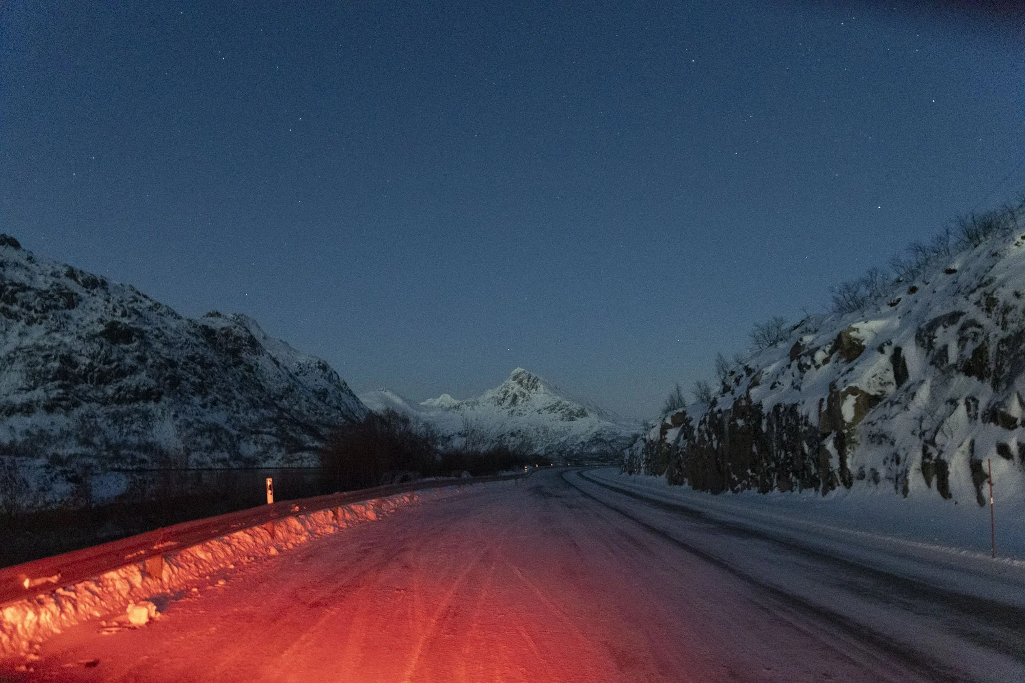 Route enneigée dans une vallée montagneuse, éclairée par les phares d'une voiture, avec un ciel étoilé au-dessus de montagnes enneigées à Tromsø