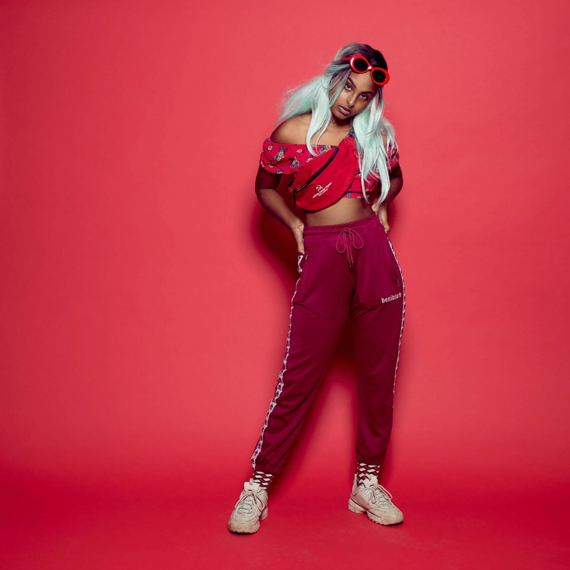 Jeune femme posant contre un mur rose, portant des vêtements confortables de sport, une veste rouge à fleurs, un pantalon de jogging bordeaux, des baskets blanches et des lunettes de soleil rouges.