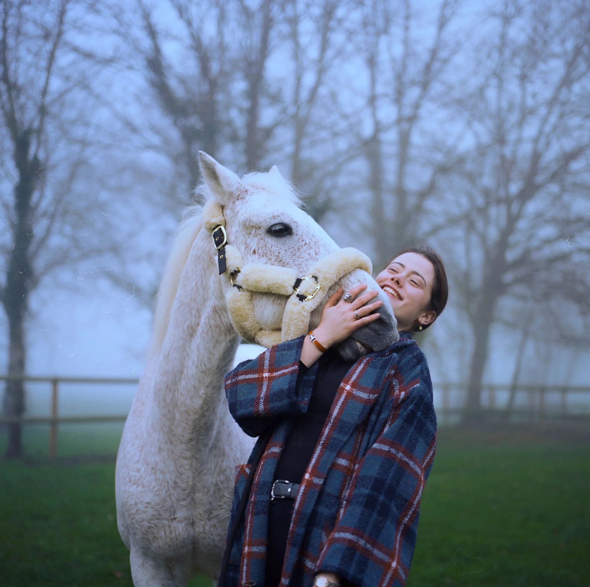 Une femme souriante qui serre un cheval blanc dans ses bras à l'extérieur, avec des arbres en arrière-plan.