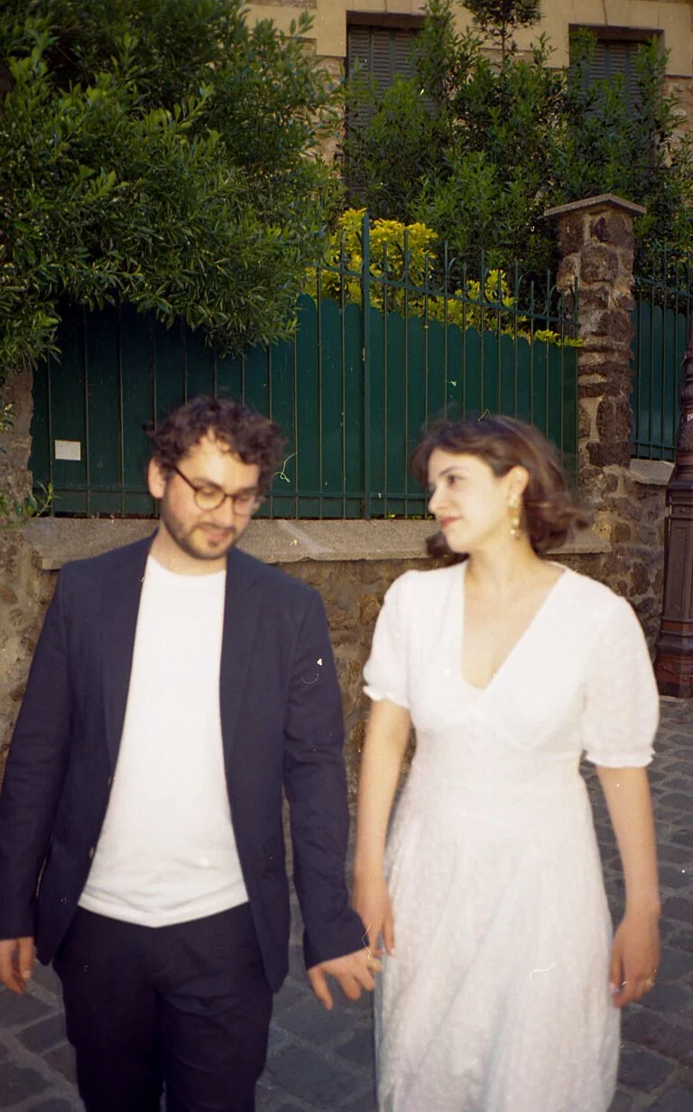 Un homme et une femme marchant main dans la main dans une rue pavée, la femme porte une robe blanche et des boucles d'oreilles, l'homme porte une veste noire et des lunettes.