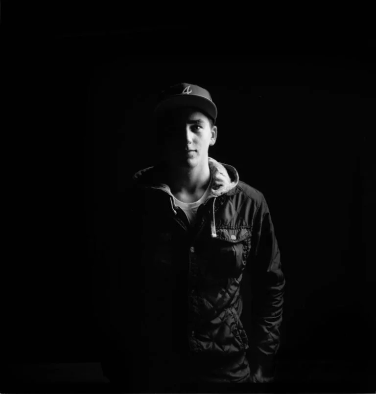 Joseph Garbaccio portant une casquette, un sweat à capuche et une veste, se tient dans un environnement sombre. La moitié de son visage est dans l'ombre, créant un contraste intense en noir et blanc.