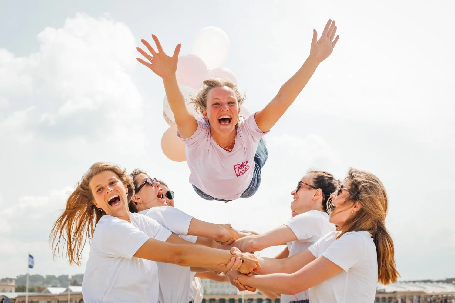 Un groupe de femmes souriantes en blanc, soulevant une femme blonde qui court dans les airs, avec des ballons roses et blancs en arrière-plan, sous un ciel bleu avec des nuages.